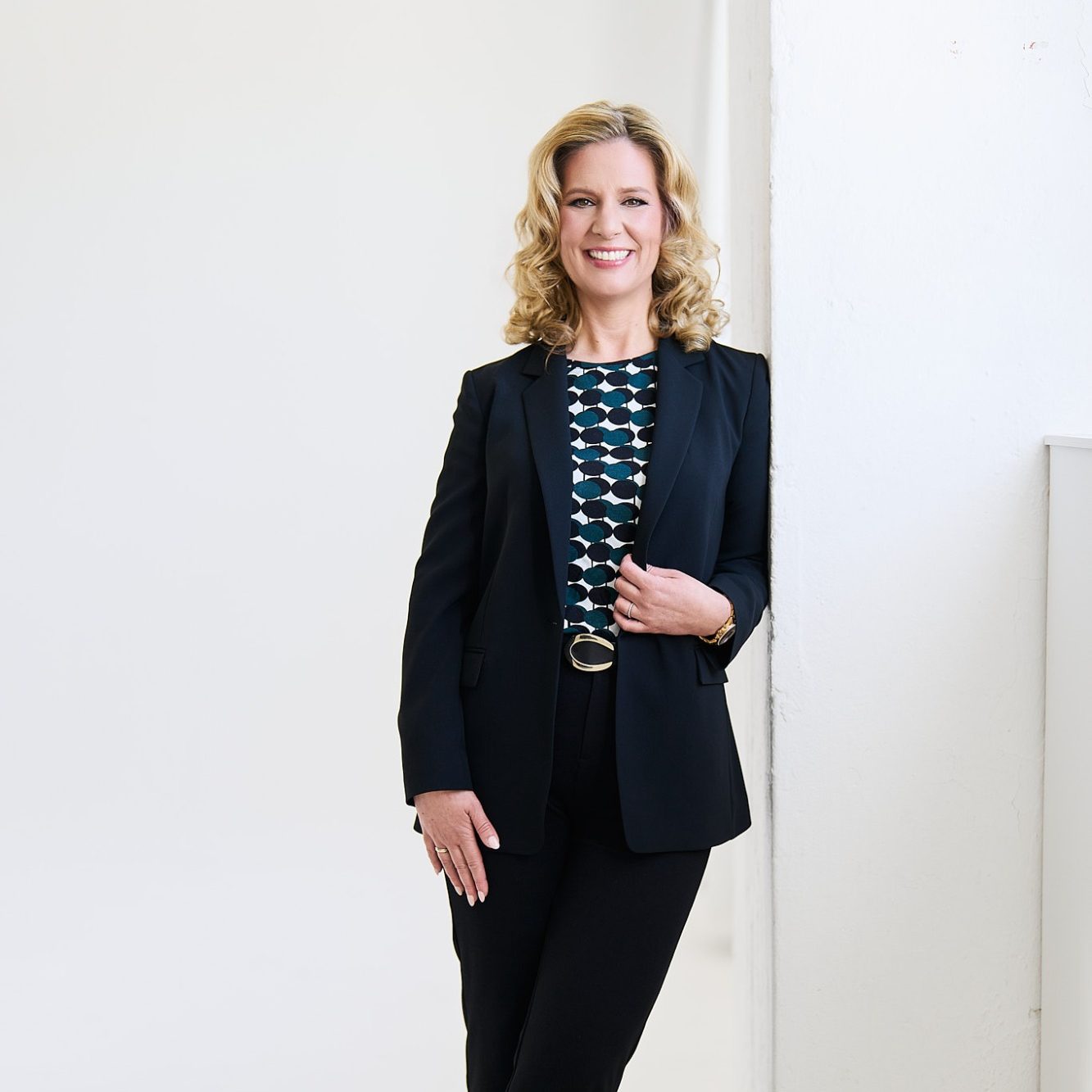 Natascha Enders in einem schwarzen Anzug, lächelnd, mit blauer und grüner Bluse im modernen Raum.