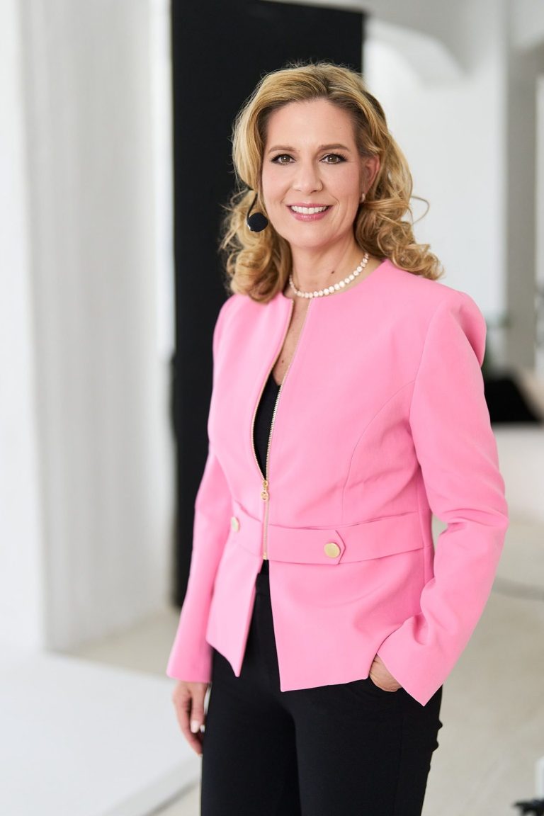 Natascha Enders – Moderation & Keynote Female Finance Natascha Enders in pinker Jacke, lächelt vor neutralem Hintergrund.